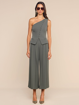 A woman wearing sunglasses and the Zinnia One-Shoulder Top, styled with wide-leg pants and heels, stands against a beige background.