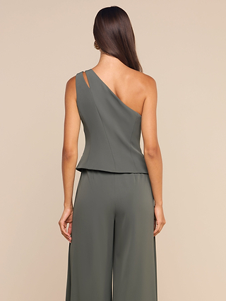 A woman with long brown hair wears the Zinnia One-Shoulder Top in ash olive, featuring sculptural tailoring. She stands with her back to the camera against a plain beige background.