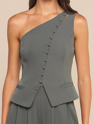 A woman wears the Zinnia One-Shoulder Top in gray, featuring sculptural tailoring, side buttons, and two front pockets, paired with a matching skirt.