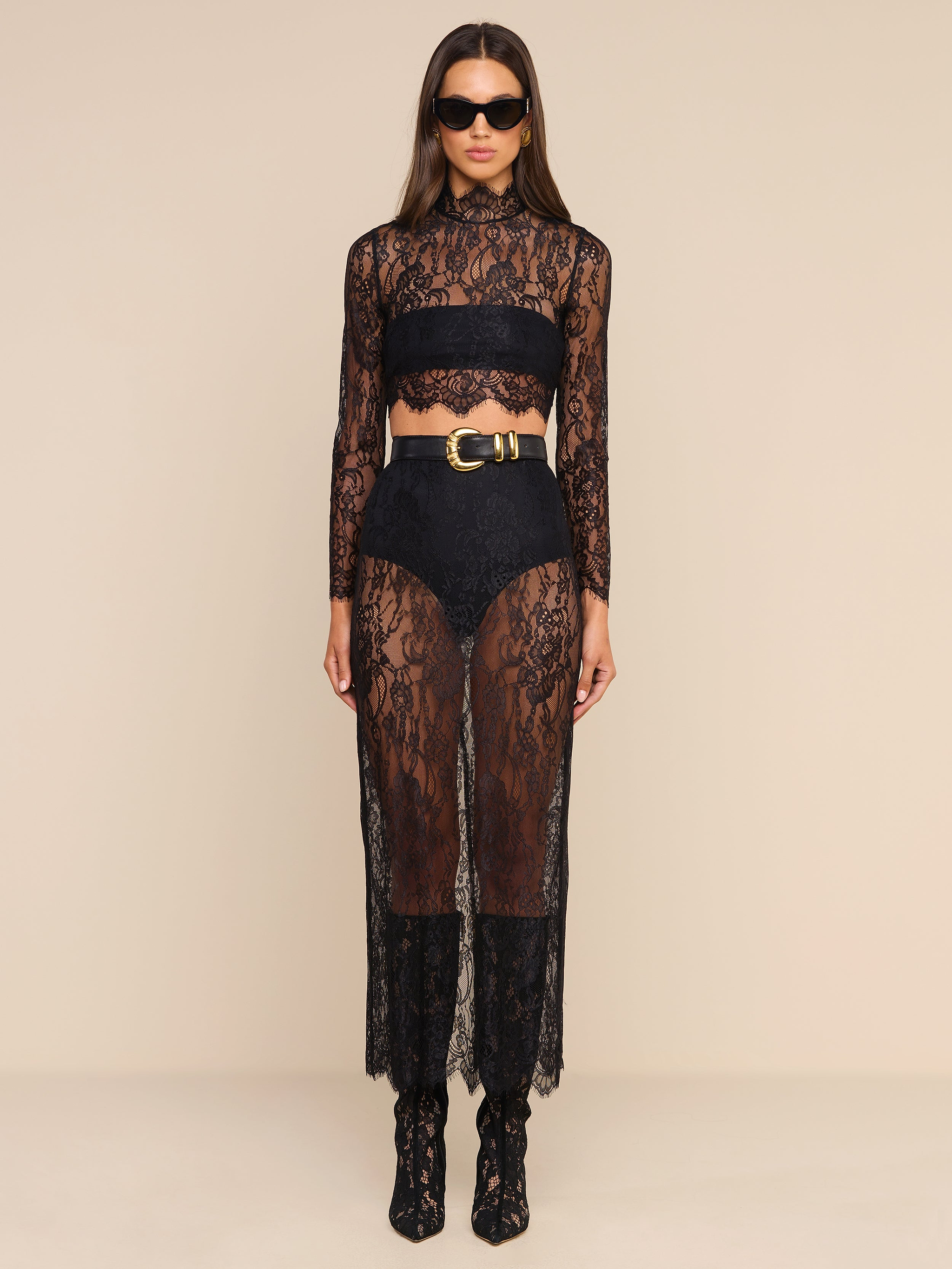 A woman wears the Henriette Cropped Lace Top—a sheer black lace long-sleeved piece with a scalloped hem—styled with high-waisted shorts, a wide belt, black ankle boots, and sunglasses as she stands against a plain beige background.