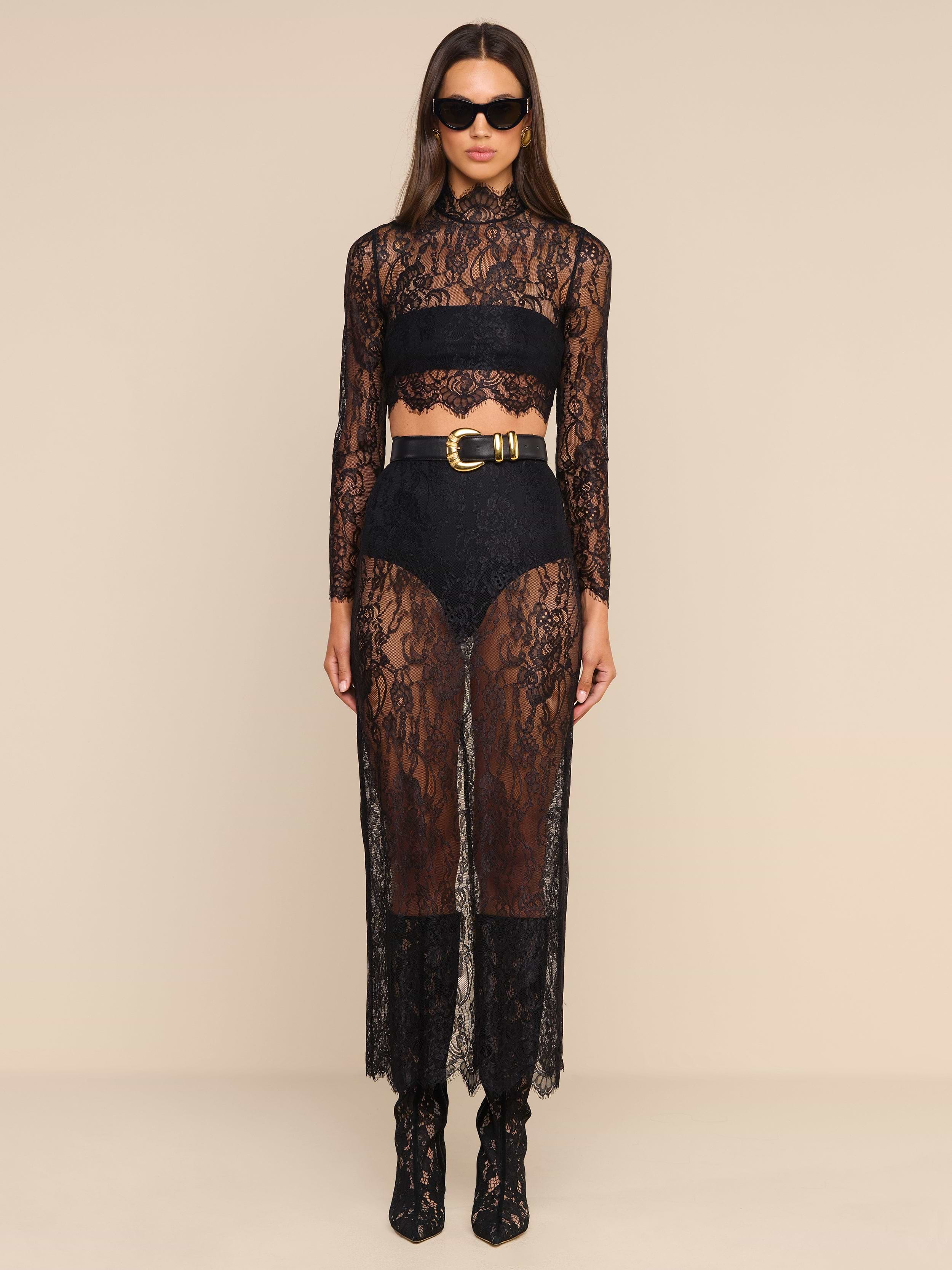 A woman wears the Henriette Cropped Lace Top—a sheer black lace long-sleeved piece with a scalloped hem—styled with high-waisted shorts, a wide belt, black ankle boots, and sunglasses as she stands against a plain beige background.