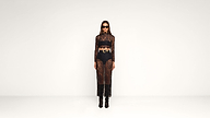 A woman stands against a plain white background wearing a sheer black lace dress, black bandeau, high-waisted shorts, black boots, large belt, and dark sunglasses.