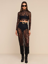 Dressed in the Henriette Cropped Lace Top with its scalloped hem, she pairs the look with high-waisted shorts, matching sheer pants, a coordinating long-sleeved top, and a belt at the waist. Sunglasses add to her style as she stands facing forward against a plain beige background.