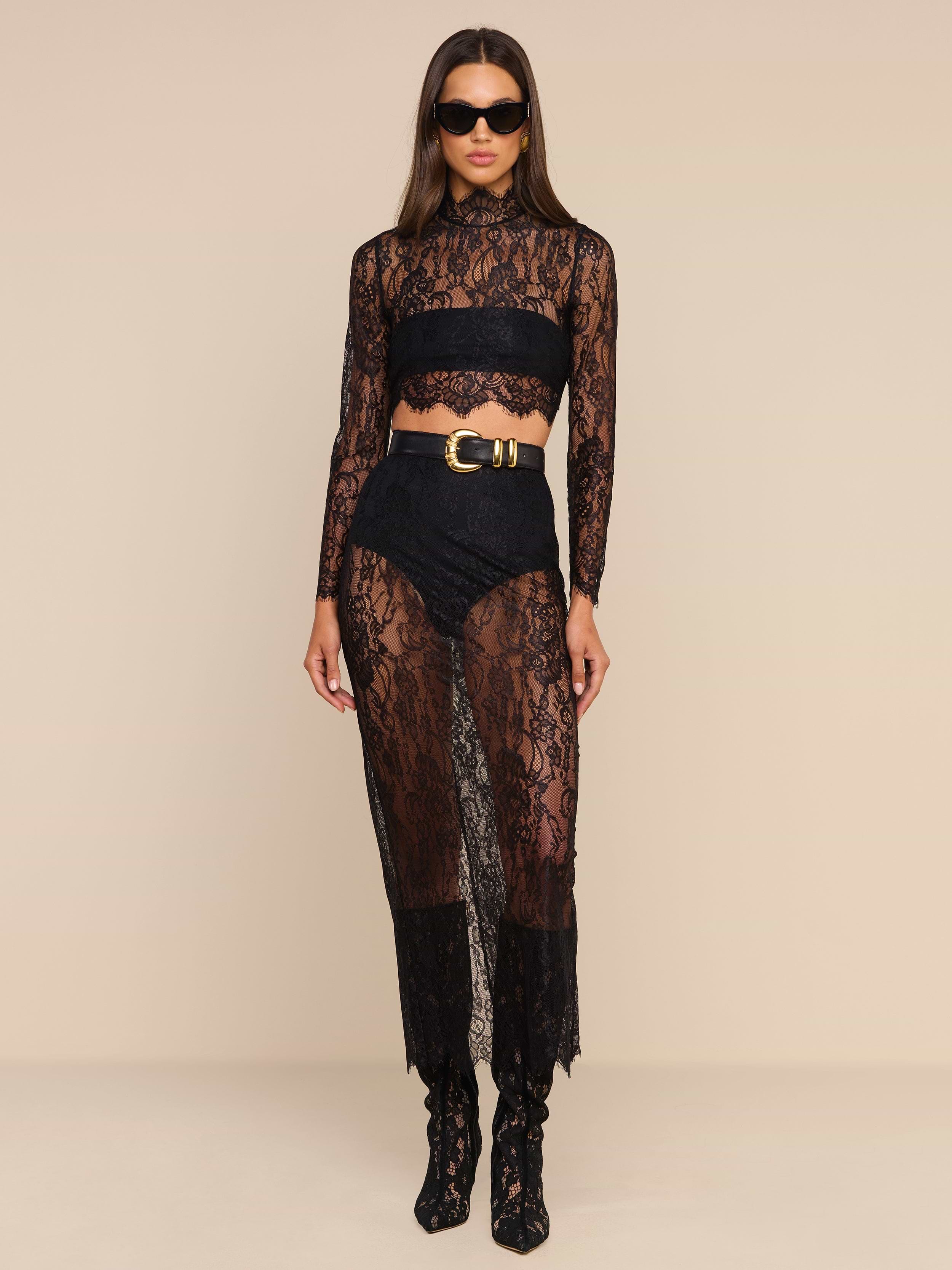 Dressed in the Henriette Cropped Lace Top with its scalloped hem, she pairs the look with high-waisted shorts, matching sheer pants, a coordinating long-sleeved top, and a belt at the waist. Sunglasses add to her style as she stands facing forward against a plain beige background.