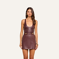 A woman stands facing forward, wearing the Tullia Scoopneck Tank in a mahogany brown vegan leather finish, styled as a sleeveless mini dress with gold buttons at the waist, and accessorized with hoop earrings and bangles.