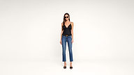 Woman wearing black tank top, blue jeans, black high heel sandals, and sunglasses stands against a plain white background, facing forward with her arms at her sides.