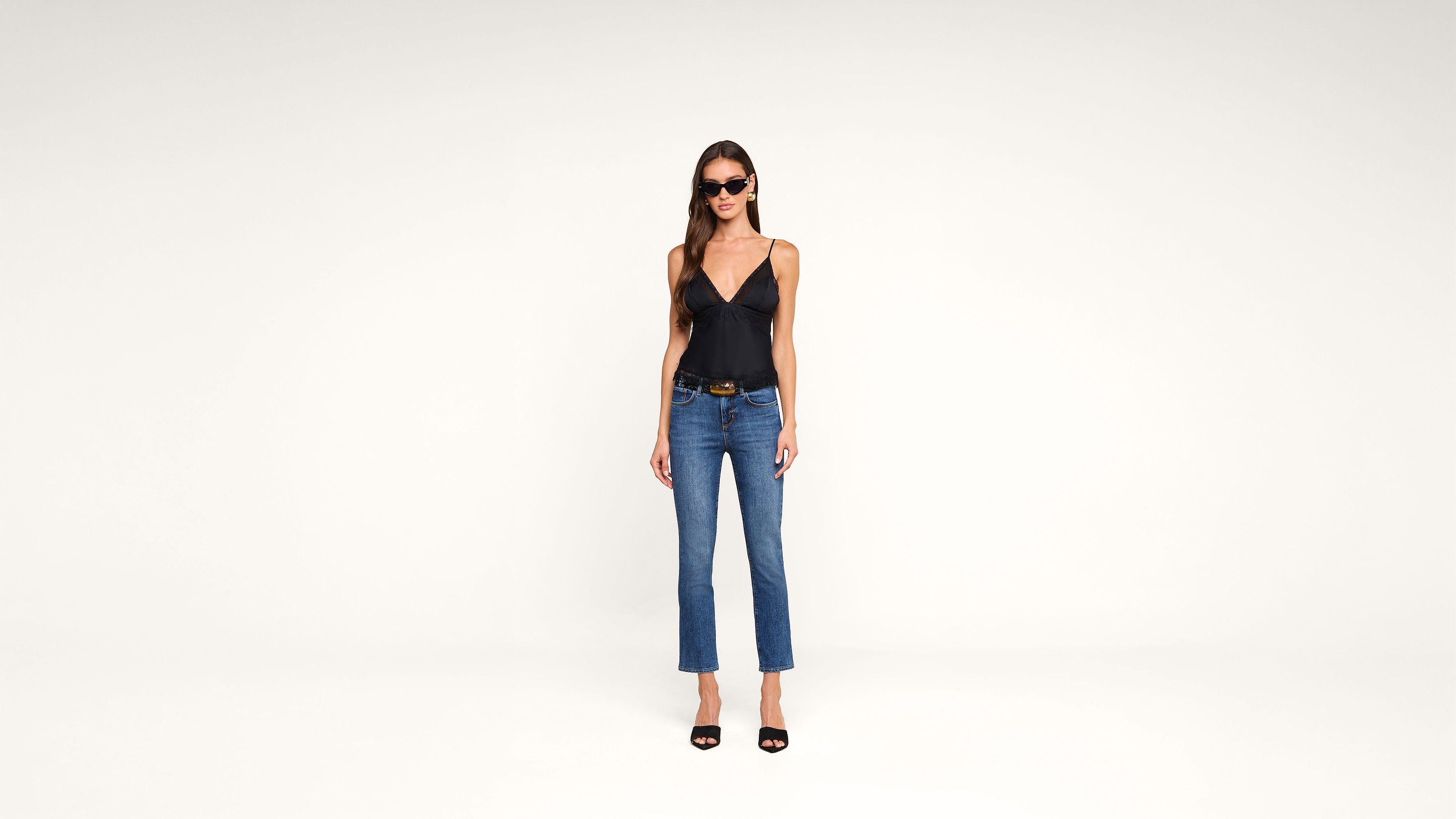 Woman wearing black tank top, blue jeans, black high heel sandals, and sunglasses stands against a plain white background, facing forward with her arms at her sides.