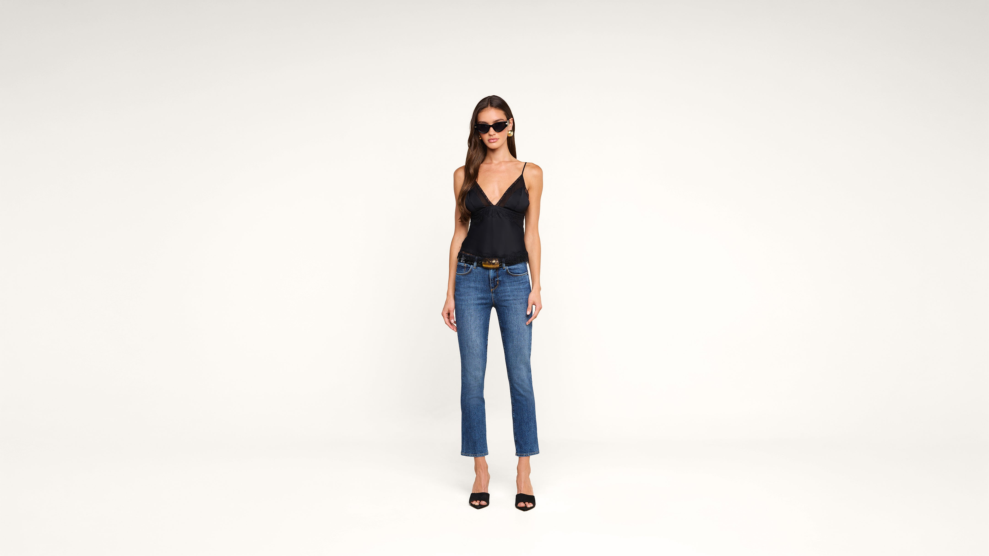 Woman wearing black tank top, blue jeans, black high heel sandals, and sunglasses stands against a plain white background, facing forward with her arms at her sides.