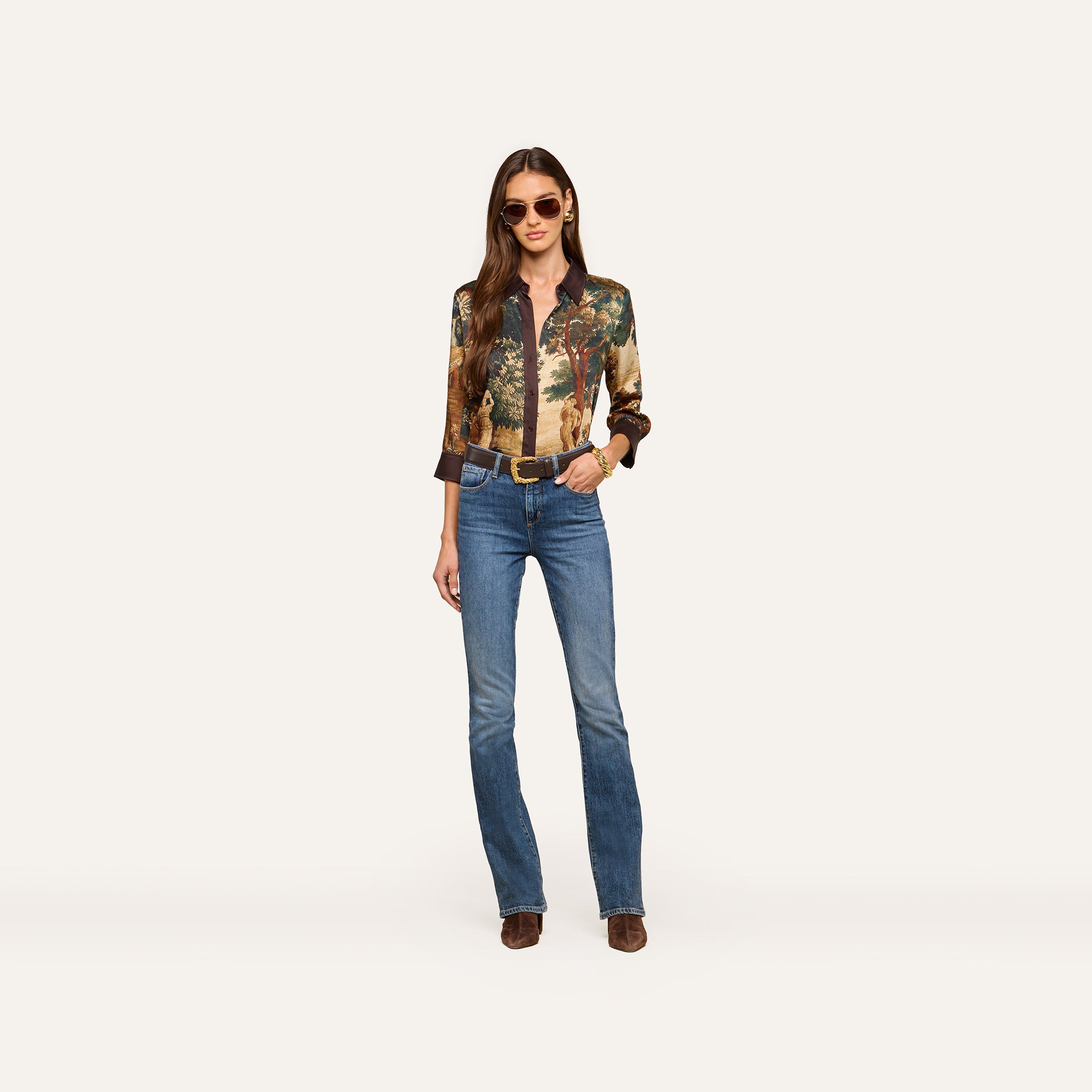 A woman stands against a plain light background, looking confidently at the camera with her hands in her pockets. She is wearing sunglasses, blue jeans, brown shoes, and the Eulalie Blouse.