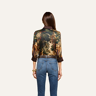 A woman with long brown hair, seen from behind, wears the Eulalie Blouse, featuring a tapestry print with a nature scene, paired with blue jeans. She stands against a plain light background.