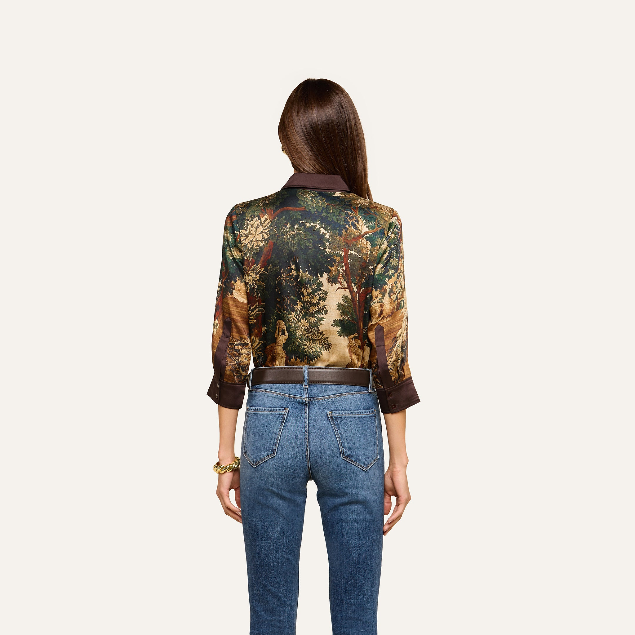 A woman with long brown hair, seen from behind, wears the Eulalie Blouse, featuring a tapestry print with a nature scene, paired with blue jeans. She stands against a plain light background.