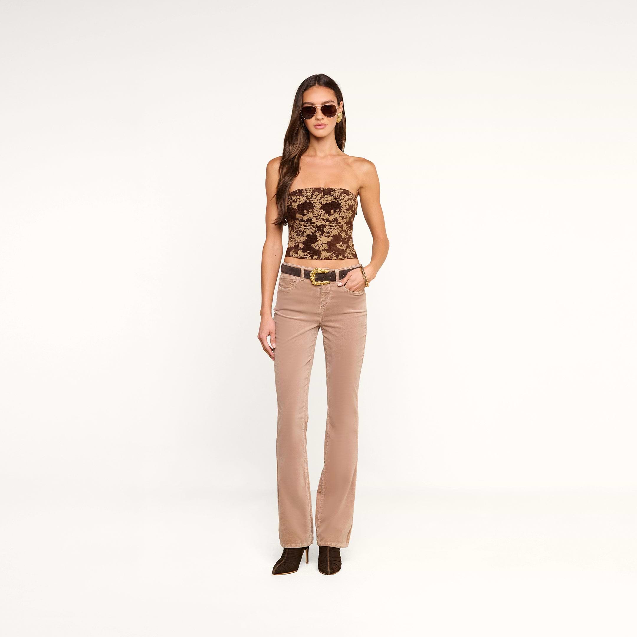 A fashion-forward woman wearing sunglasses, the Paulette Corset Top in a floral jacquard pattern, beige flared pants with a belt, and brown boots stands against a plain white background.