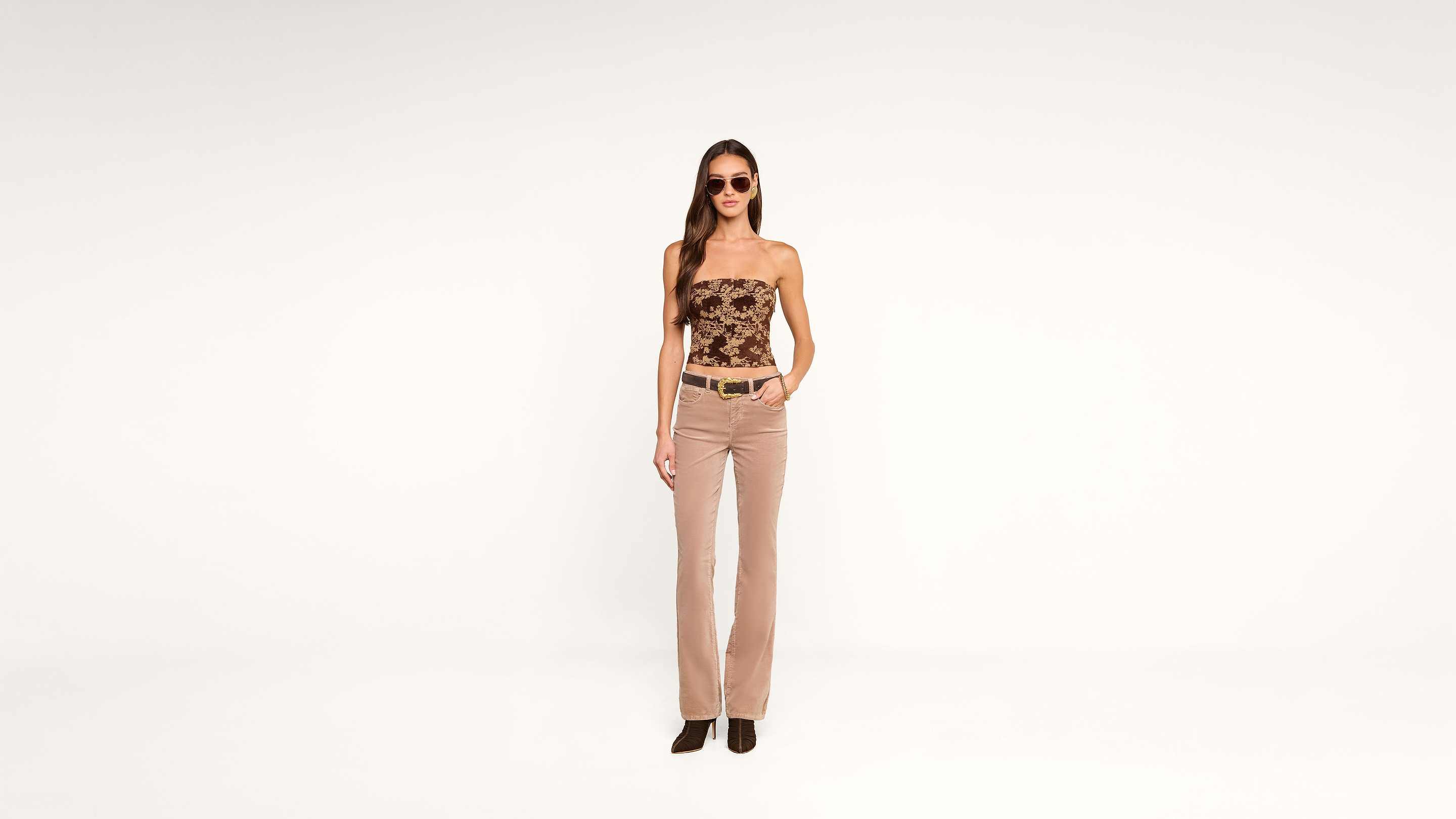 A woman wearing sunglasses, a strapless patterned top, beige pants, and black shoes stands against a plain white background with one hand in her pocket.