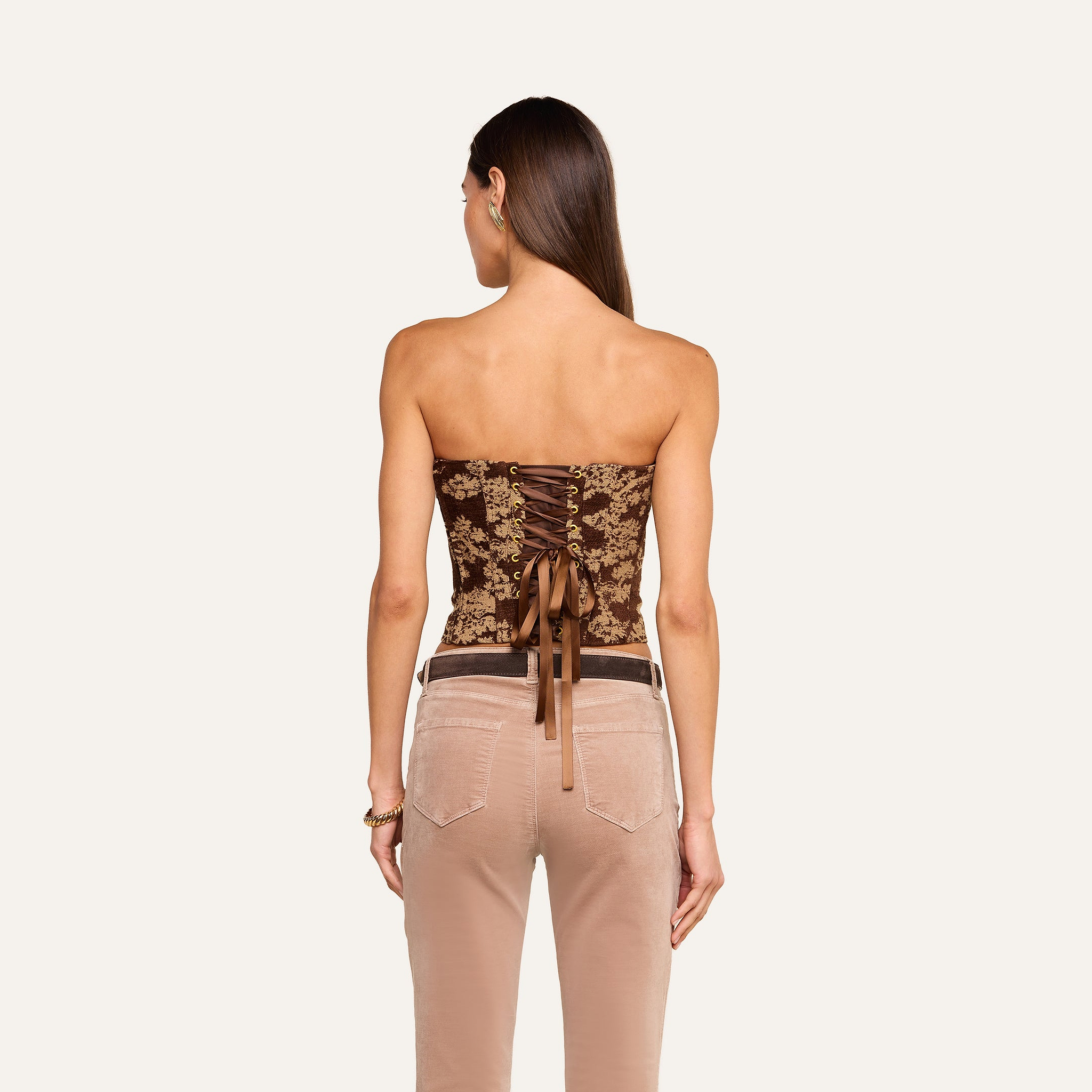 A woman with straight, loose hair faces away, wearing the Paulette Corset Top—a brown strapless piece featuring lace-up detailing, a bow accent, and crafted in a fashion-forward floral jacquard fabric. She pairs it with beige pants that have visible back pockets.