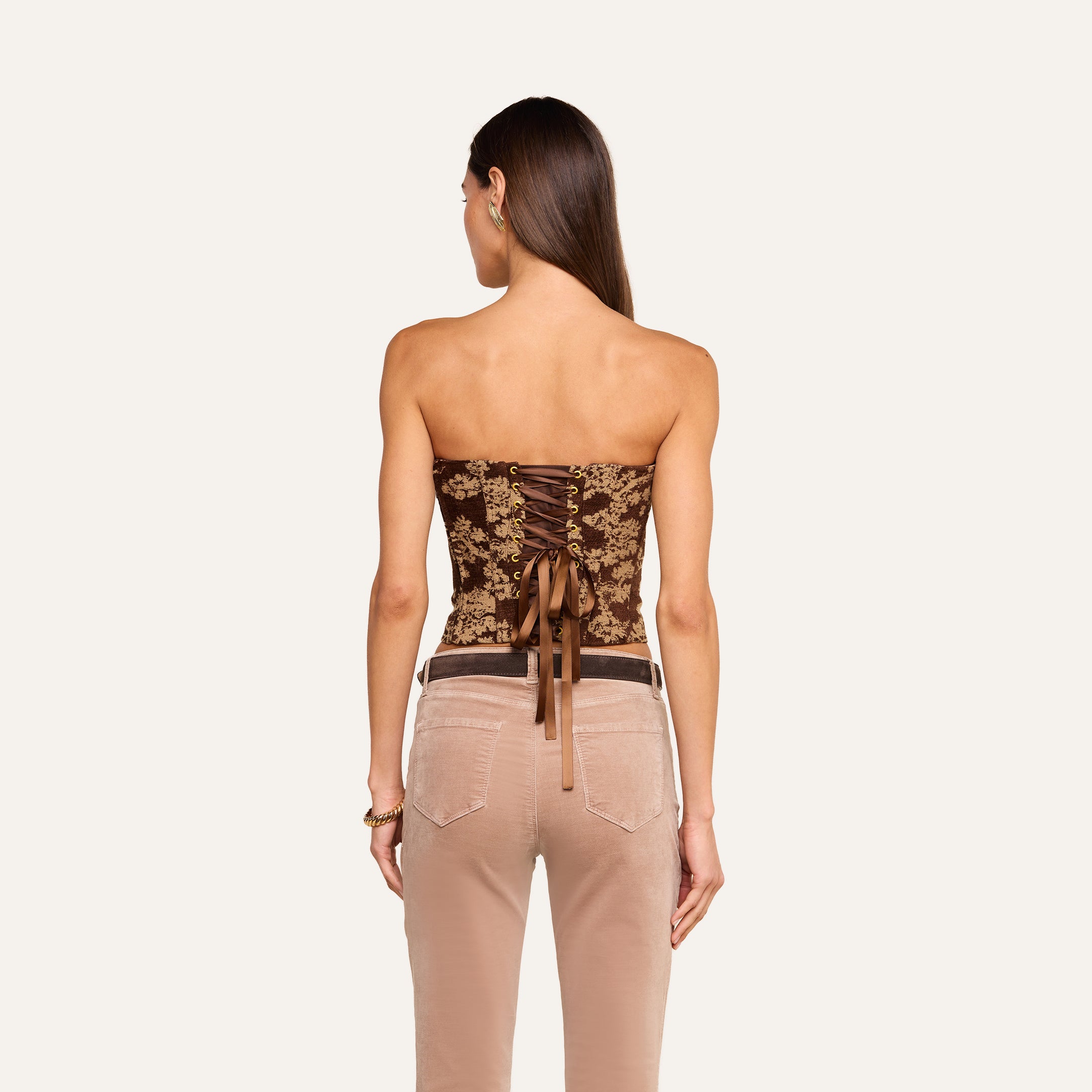 A woman with straight, loose hair faces away, wearing the Paulette Corset Top—a brown strapless piece featuring lace-up detailing, a bow accent, and crafted in a fashion-forward floral jacquard fabric. She pairs it with beige pants that have visible back pockets.