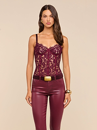 A woman stands facing forward against a beige background, wearing the Jenny Lace Bustier in burgundy paired with matching leather pants featuring a gold-buckled belt. She accessorizes her look with gold earrings and a bracelet. The intricate detailing of the Jenny Lace Bustier’s underwire design beautifully enhances her silhouette, adding an elegant touch to her ensemble.