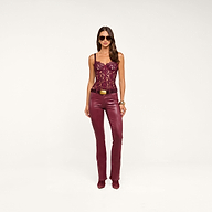 A woman wearing sunglasses, the Jenny Lace Bustier in burgundy, matching leather pants with a belt, gold bracelets, and burgundy shoes stands against a plain beige background.
