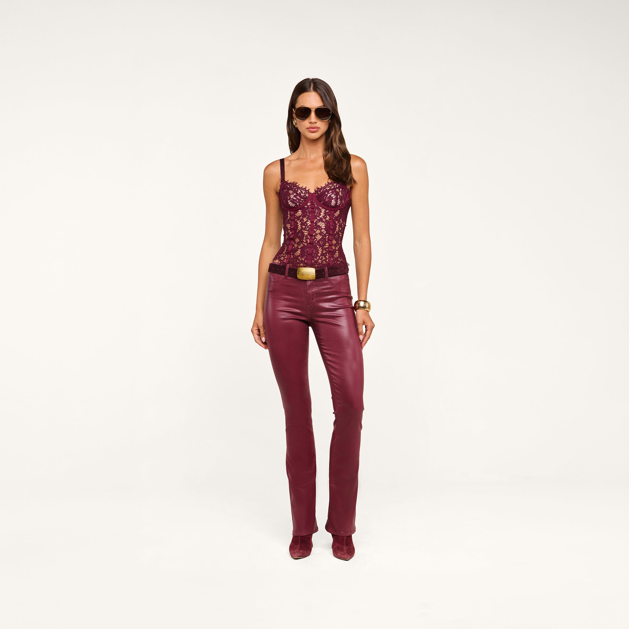 A woman wearing sunglasses, the Jenny Lace Bustier in burgundy, matching leather pants with a belt, gold bracelets, and burgundy shoes stands against a plain beige background.
