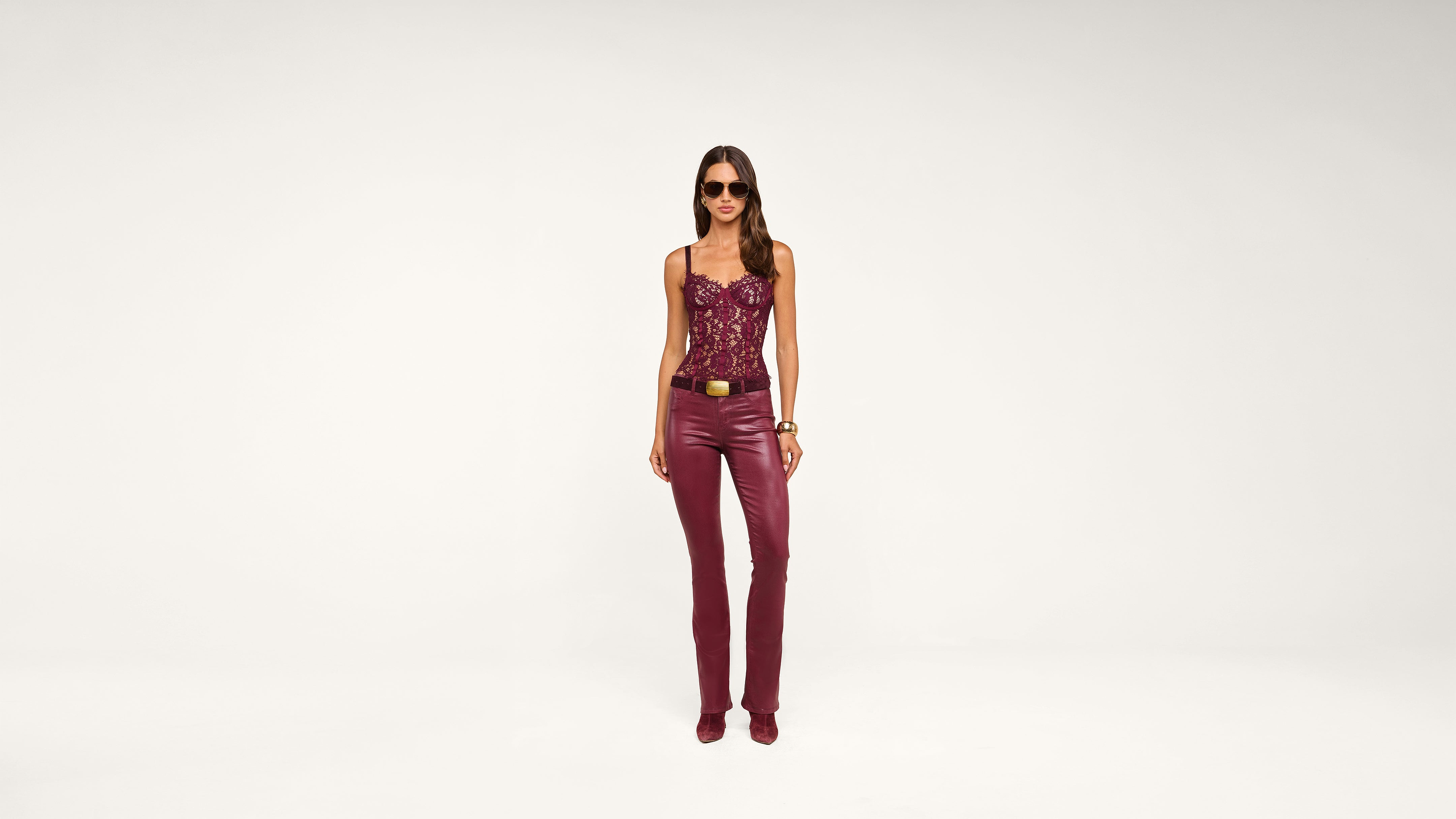 Woman in burgundy lace top and matching burgundy leather pants stands facing forward, wearing sunglasses and heels, with a neutral white background.