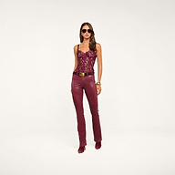 A woman wearing sunglasses, the Jenny Lace Bustier in burgundy floral lace, paired with burgundy leather belted pants and matching heeled boots, stands against a neutral background.