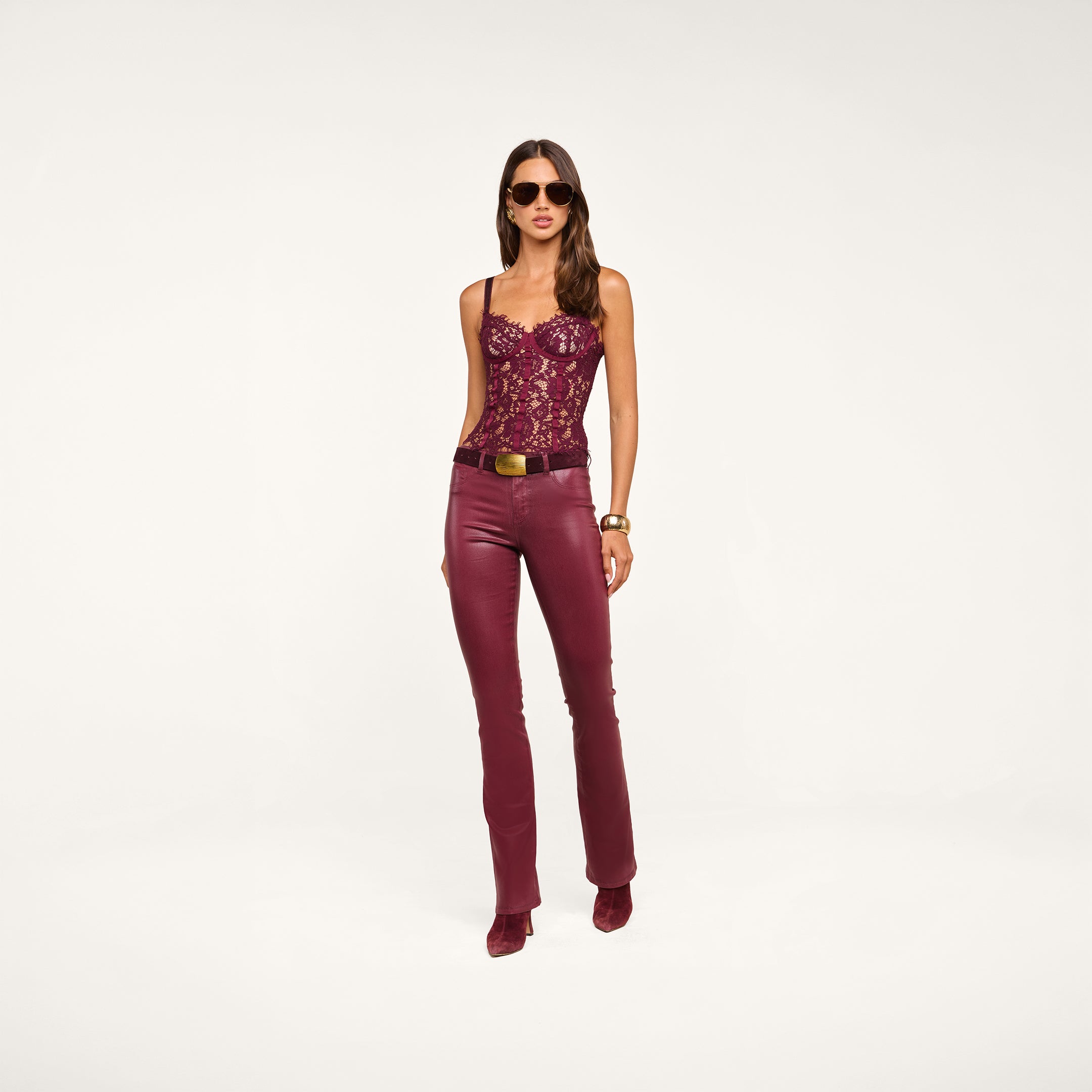 A woman wearing sunglasses, the Jenny Lace Bustier in burgundy floral lace, paired with burgundy leather belted pants and matching heeled boots, stands against a neutral background.