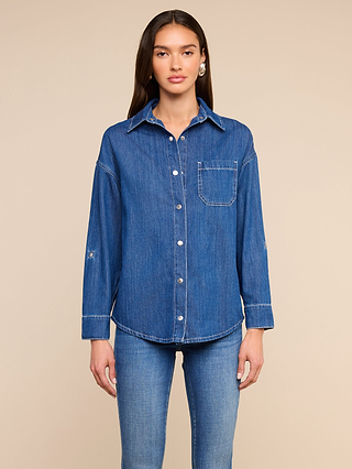 Woman standing against a beige background, wearing the Jorah Oversized Shirt—a long-sleeve, blue featherweight denim shirt featuring snap button closures and a chest pocket—paired with blue jeans. She has long, straight brown hair and is looking forward.