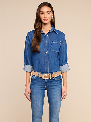A woman stands facing forward, wearing the Jorah Oversized Shirt in blue denim with rolled-up sleeves and snap button closures, paired with matching blue jeans, a beige belt, and a single large earring. The background is plain beige.