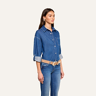 A woman wearing the Jorah Oversized Shirt with rolled-up sleeves, paired with indigo blue jeans, a beige belt, and large earrings, stands against a plain light background.