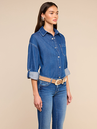 A woman stands against a beige background wearing the Jorah Oversized Shirt with rolled sleeves and snap button closures, paired with blue jeans, a tan belt, and statement earrings.
