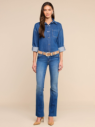 A woman stands against a beige background wearing the Jorah Oversized Shirt with rolled sleeves and snap button closures, styled with matching blue jeans, a tan belt, and brown pointed-toe heels. Her long hair is down as she faces forward with a neutral expression.