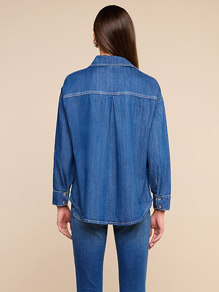 A woman with long brown hair, wearing the Jorah Oversized Shirt—an extra-roomy featherweight denim top with snap button closures—and blue jeans, stands facing away from the camera against a plain beige background.