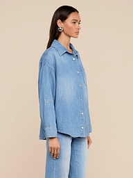 Une femme aux longs cheveux bruns se tient de profil sur un fond beige, portant la chemise Jorah Oversized Shirt - un modèle en denim ultra-léger avec des manches à pattes roulées - associée à un jean assorti.