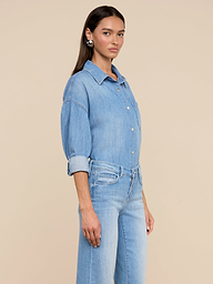 Une femme aux longs cheveux bruns porte la chemise surdimensionnée Jorah en denim bleu clair léger et un jean taille haute assorti, sur un fond beige uni.