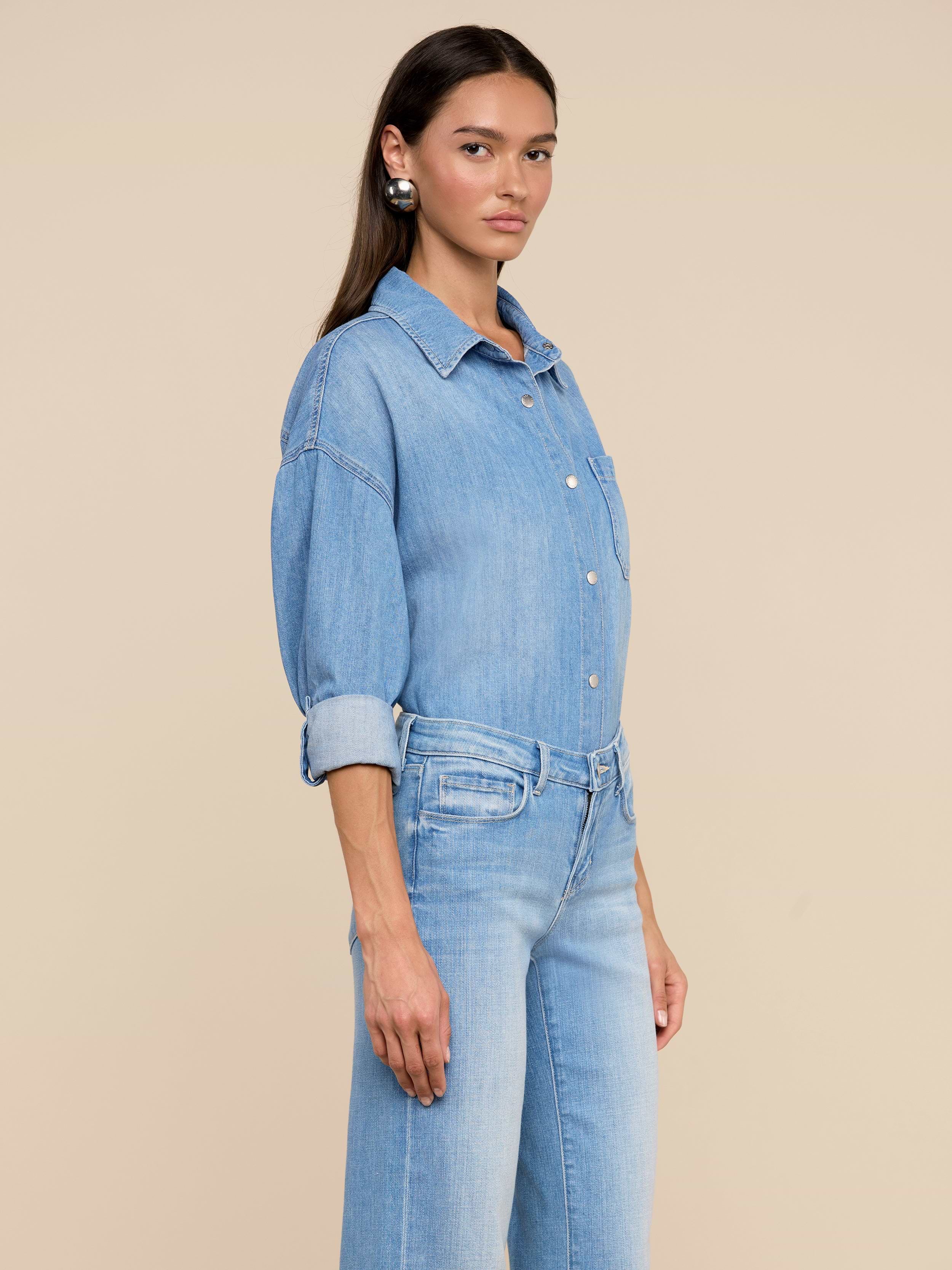 Une femme aux longs cheveux bruns porte la chemise surdimensionnée Jorah en denim bleu clair léger et un jean taille haute assorti, sur un fond beige uni.