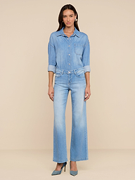 Une femme aux cheveux longs et lâchés se tient sur un fond beige. Elle porte la chemise surdimensionnée Jorah en denim poids plume, associée à un jean large bleu clair assorti et à des talons bleus. Son expression est neutre.