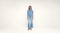 A woman stands in a minimalist studio wearing a light blue denim shirt and wide-leg jeans with teal heels. She faces forward with her hands by her sides. The background is plain and off-white.