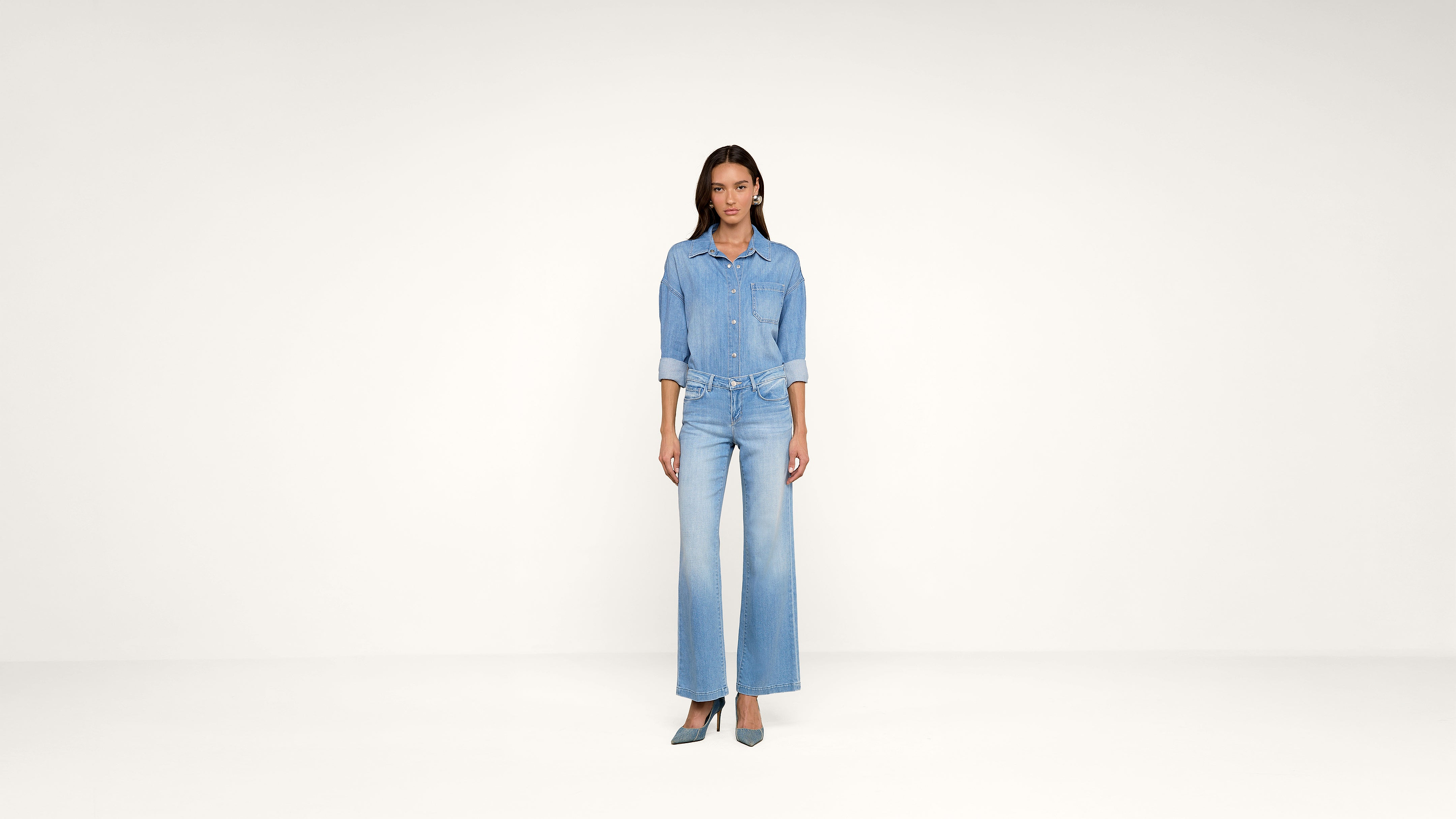 A woman stands in a minimalist studio wearing a light blue denim shirt and wide-leg jeans with teal heels. She faces forward with her hands by her sides. The background is plain and off-white.