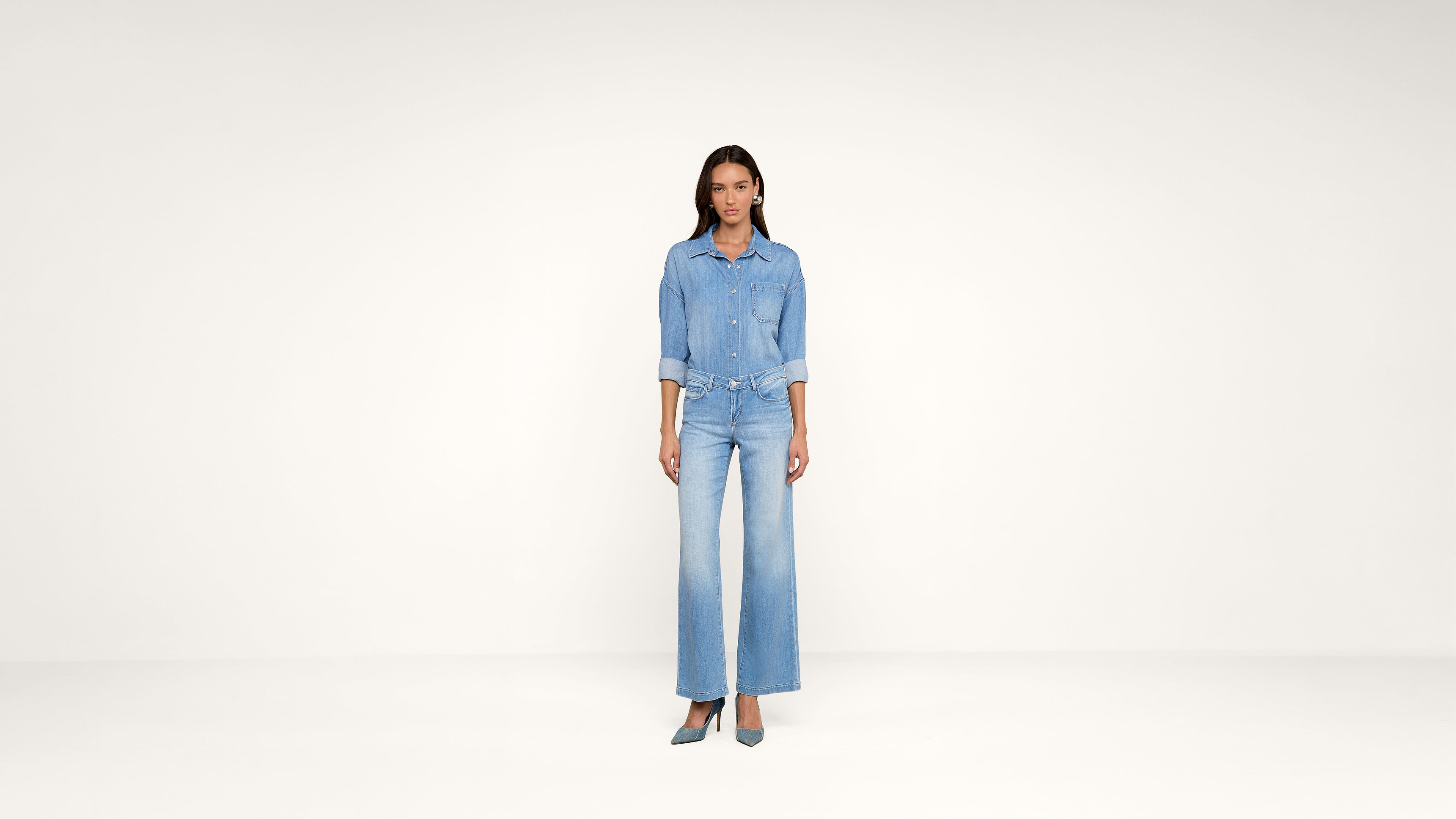 A woman stands in a minimalist studio wearing a light blue denim shirt and wide-leg jeans with teal heels. She faces forward with her hands by her sides. The background is plain and off-white.