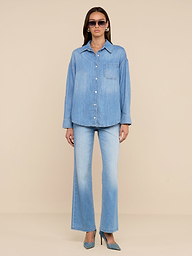 Une femme se tient sur un fond beige, portant des lunettes de soleil, des chaussures bleues à talons hauts, un jean évasé en denim bleu clair et la chemise surdimensionnée Jorah - une chemise en denim poids plume avec une poche sur le devant et des manches à patte de boutonnage.