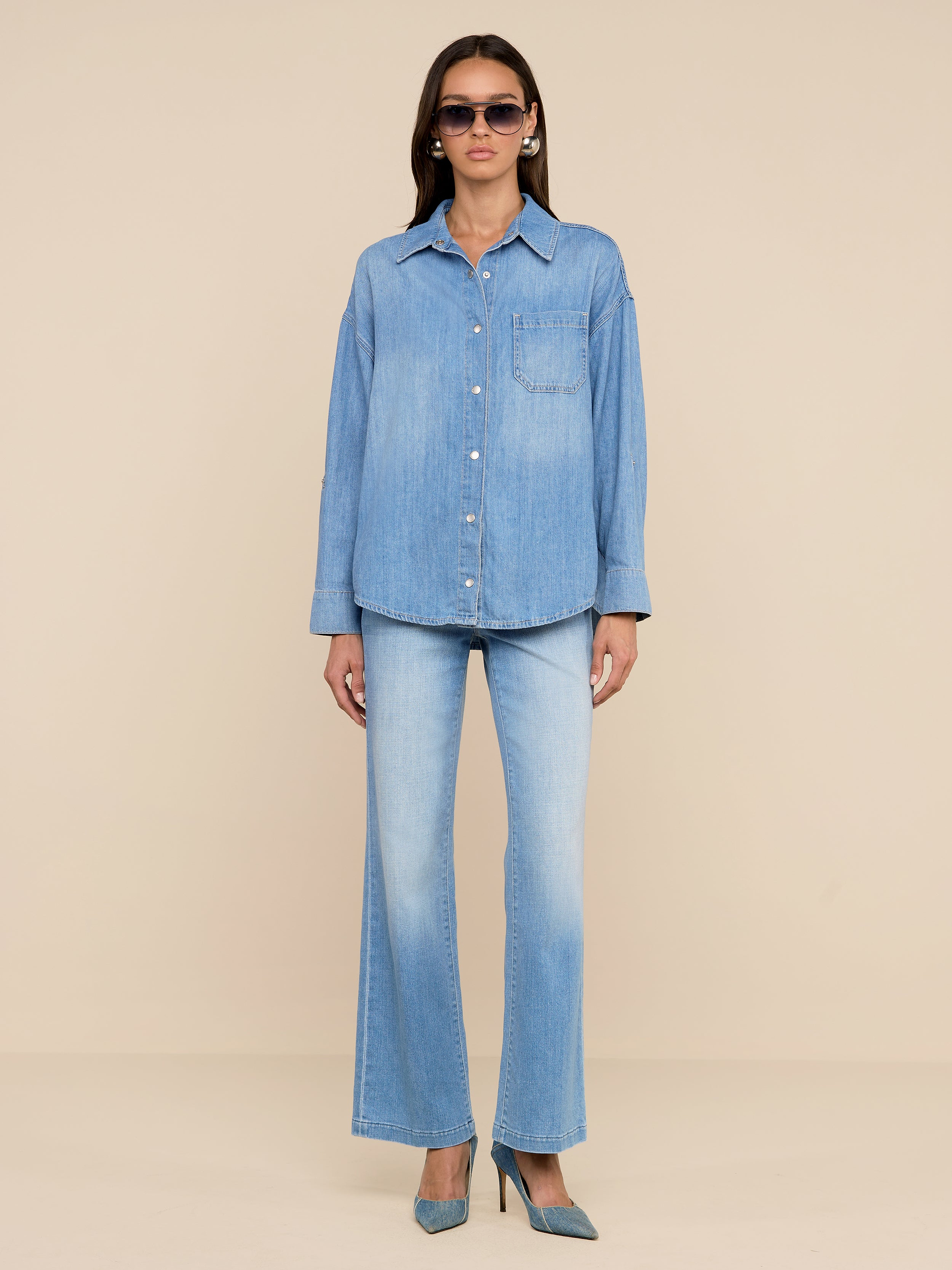 Une femme se tient sur un fond beige, portant des lunettes de soleil, des chaussures bleues à talons hauts, un jean évasé en denim bleu clair et la chemise surdimensionnée Jorah - une chemise en denim poids plume avec une poche sur le devant et des manches à patte de boutonnage.