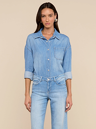 Une femme aux longs cheveux bruns se tient sur un fond beige, vêtue de la chemise surdimensionnée Jorah - un haut en denim poids plume avec des manches à pattes retroussées - associée à un jean bleu clair assorti, et regardant directement la caméra.