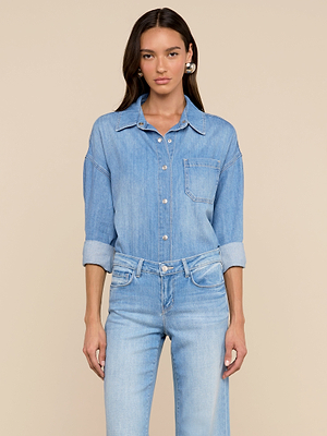 A woman with long dark hair stands against a beige backdrop, wearing the Jorah Oversized Shirt— a featherweight denim top with roll-tab sleeves—paired with matching light blue jeans, looking directly at the camera.