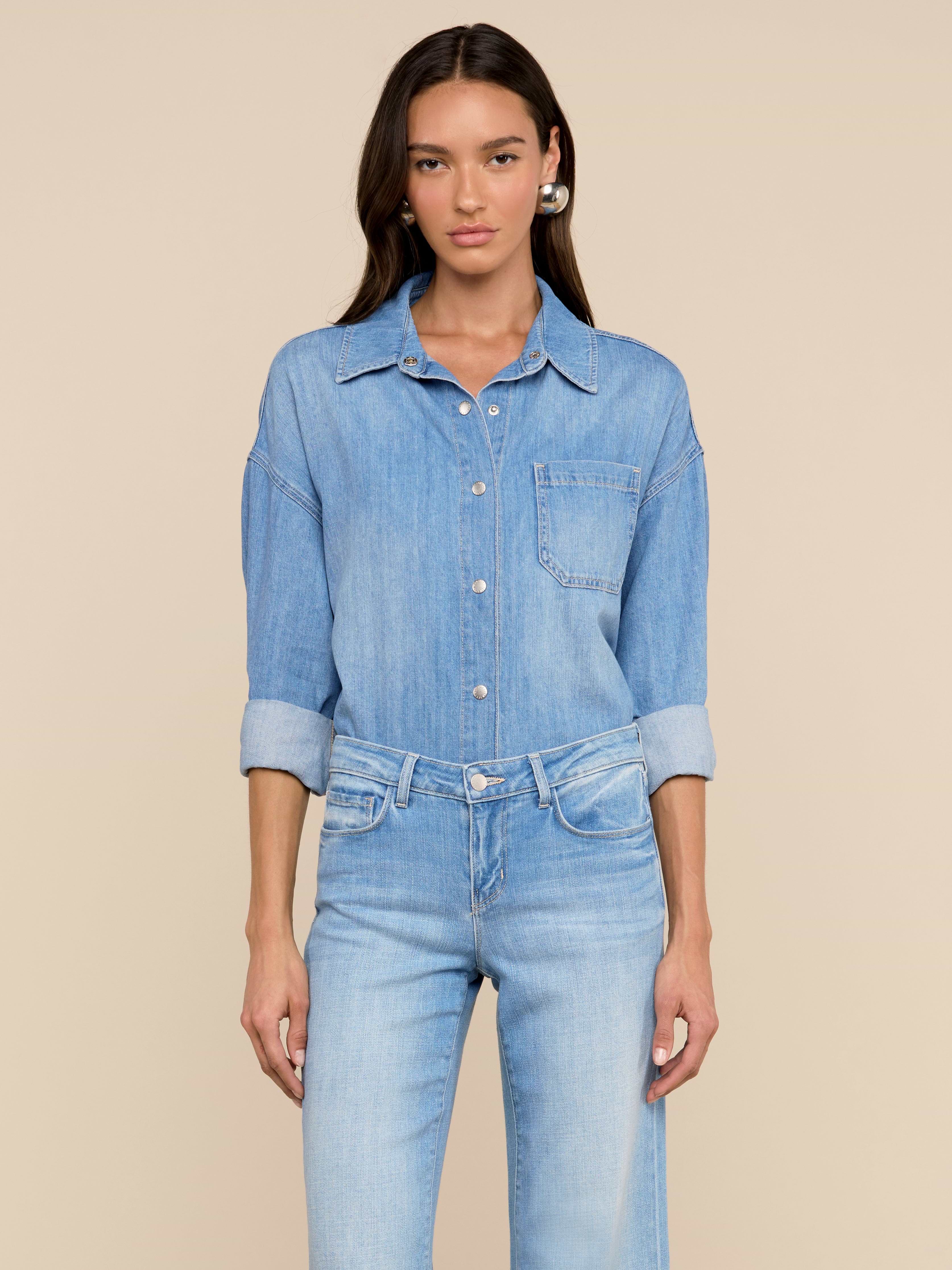 Une femme aux longs cheveux bruns se tient sur un fond beige, vêtue de la chemise surdimensionnée Jorah - un haut en denim poids plume avec des manches à pattes retroussées - associée à un jean bleu clair assorti, et regardant directement la caméra.