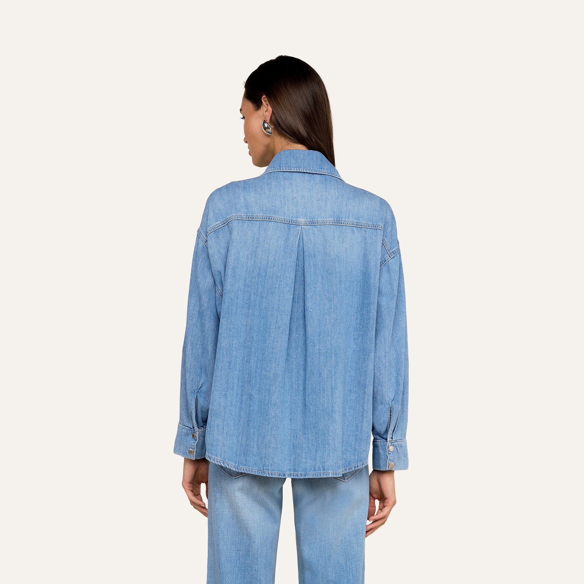 Une femme aux longs cheveux bruns est de face, portant la chemise surdimensionnée Jorah en denim poids plume bleu clair avec des manches à boutons roulés et un jean assorti, sur un fond beige uni.