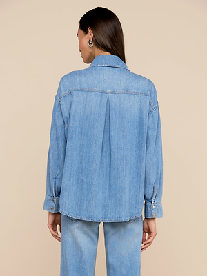 A woman with long brown hair faces away, wearing the Jorah Oversized Shirt in light blue featherweight denim with roll-tab sleeves and matching jeans, set against a plain beige background.