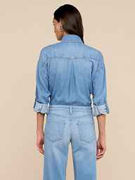 Une femme regarde au loin, portant la chemise surdimensionnée Jorah - un haut en denim ultra-léger avec des manches à pattes roulées - associée à un jean taille haute assorti sur un fond beige uni.