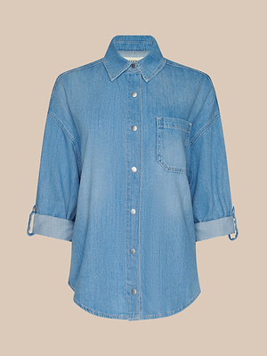 La chemise surdimensionnée Jorah en denim poids plume bleu clair présente des manches à patte de boutonnage, une poche poitrine gauche et un col pointu, le tout sur fond beige uni.