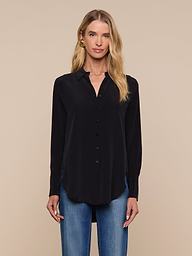 A woman stands facing forward against a plain light background, wearing the Babette Silk Blouse— a long-sleeve black crepe de chine top with a relaxed fit—paired with blue jeans.
