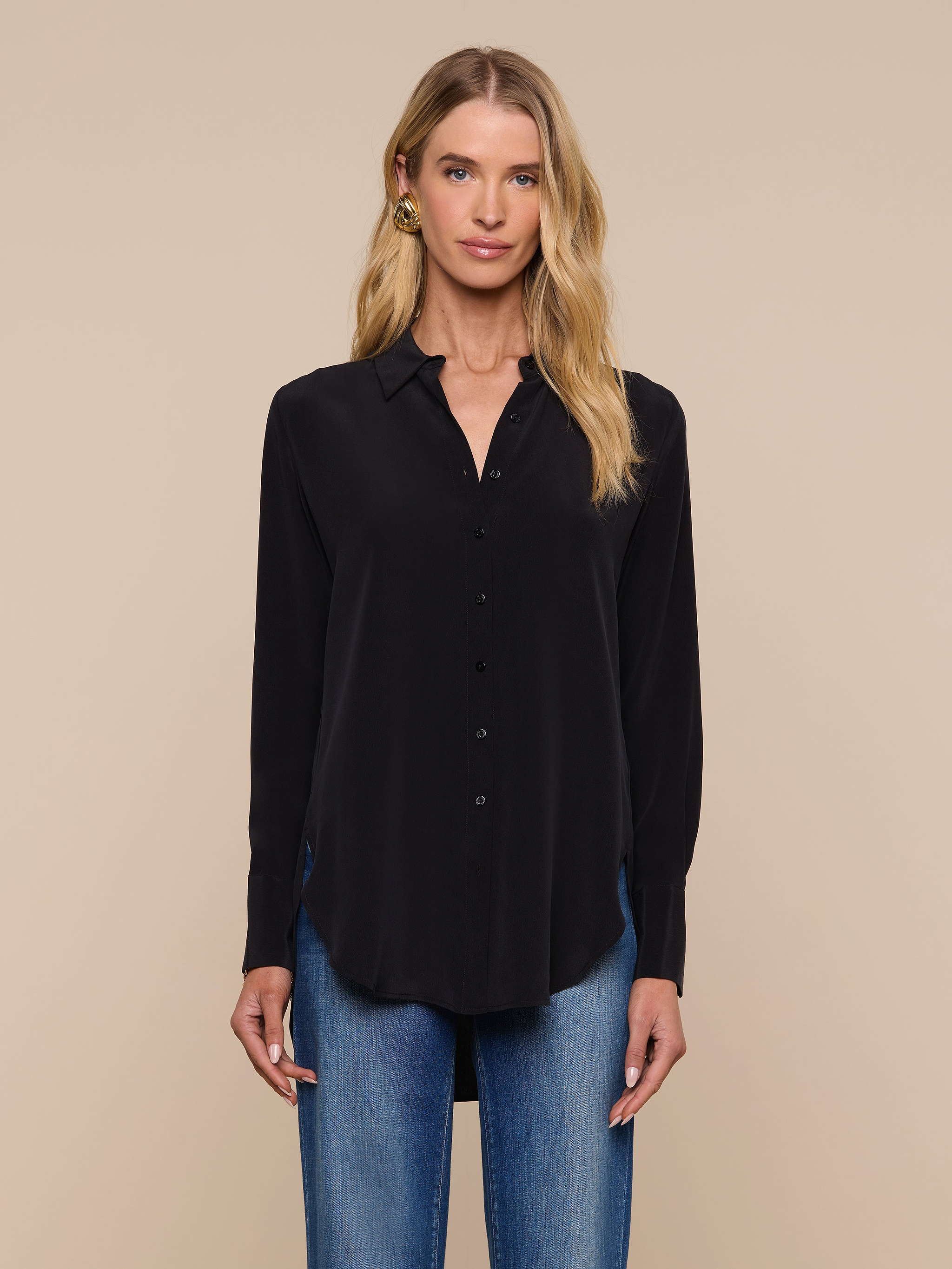 A woman stands facing forward against a plain light background, wearing the Babette Silk Blouse— a long-sleeve black crepe de chine top with a relaxed fit—paired with blue jeans.