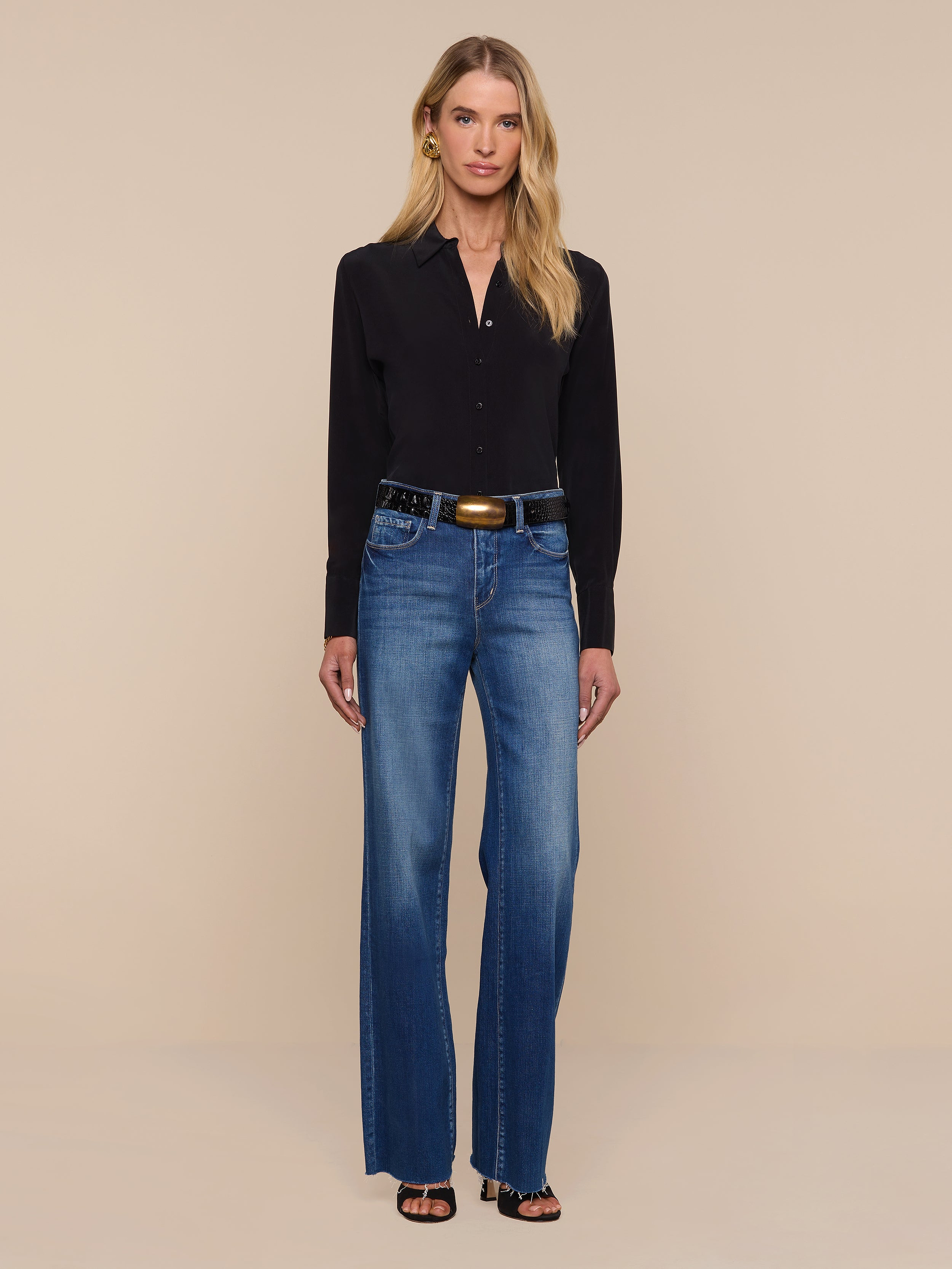 A woman stands against a plain white background, wearing the Babette Silk Blouse in black with long sleeves and a relaxed fit, paired with blue high-waisted wide-leg jeans, a black belt with a gold buckle, and black open-toe heels.